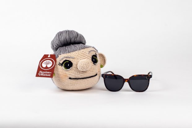 A crocheted woman’s head on a white background. Her grey hair, pulled back into a bun, is made of strands of yarn. She has a prominent nose, big green eyes and a smiling mouth, and she is wearing green earrings. The object has a red label on it. To its right there is a pair of sunglasses.