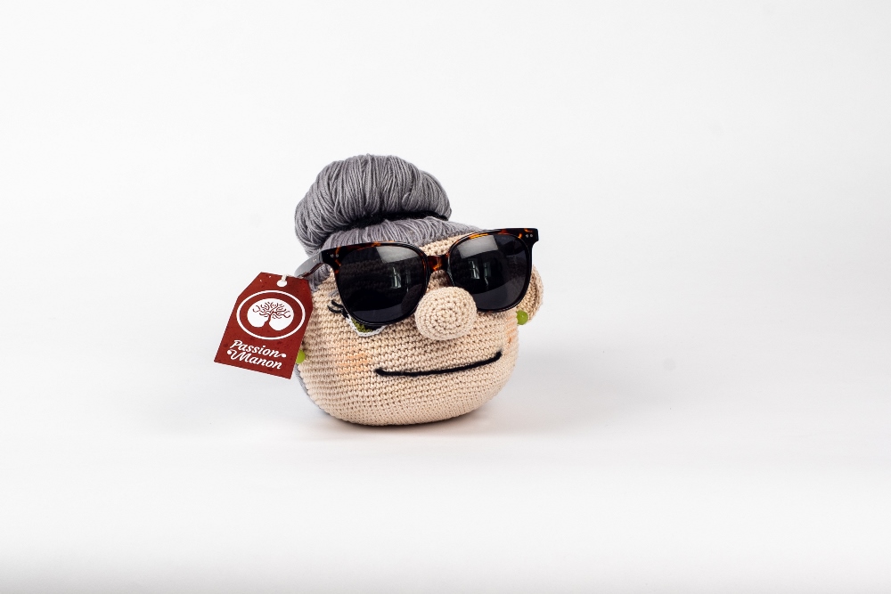 A crocheted woman’s head on a white background. Her grey hair, pulled back into a bun, is made of strands of yarn. She has a smiling mouth and a prominent nose upon which is perched a pair of sunglasses, and she is wearing green earrings. A red label is attached to the object.