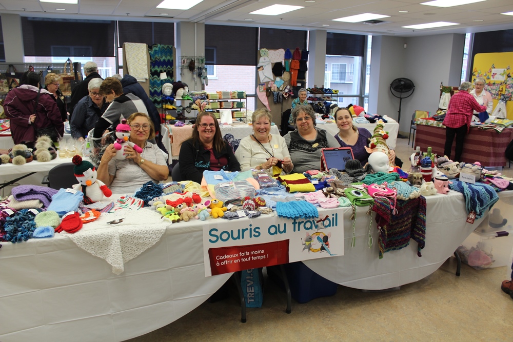 Five women sit behind a long table at the 2019 exhibition and sale, with some of their textile creations spread out in front of them. A small sign attached to the front of the table reads “Souris au travail” (“Mice at work”).