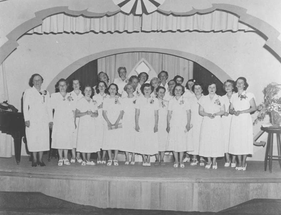 A black and white photograph of around twenty women in white wearing corsages, standing together on a stage. A piano can be seen to their left and a bouquet of flowers on a stool to their right.