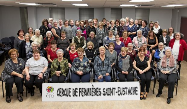 Several dozens of Fermières are gathered in a large hall for a group photograph. In front of the group is a white banner showing the logo of the Cercles de Fermières du Québec with the words “Cercle de Fermières Saint-Eustache.”