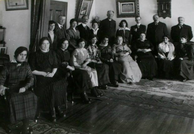 Fourteen women in long skirts or dresses and six men in suits are in the corner of a room. The women in the front row are all seated. The men and women in the back row are standing.