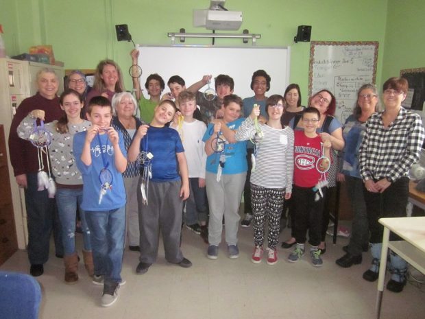 A number of children and a few smiling adults are gathered in two rows in front of a white board in a classroom. Each of the children is holding a dream catcher.