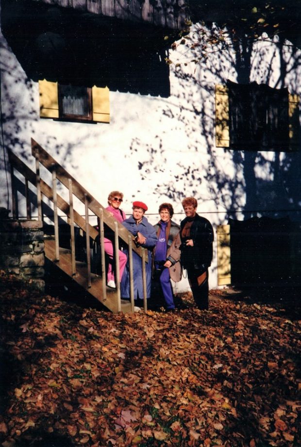 Four women are standing together outside at the bottom of a staircase in front of a white building with yellow shutters on the windows. In front of them is a carpet of dead leaves.