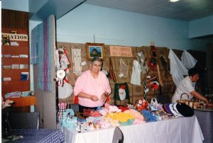A woman in a pink sweater stands behind a table with various textile creations spread out on it. More pieces hang on a board behind her.