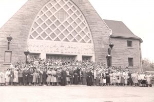 A sepia photograph of several dozen women and a few men gathered in front of a church. They are all looking straight ahead.