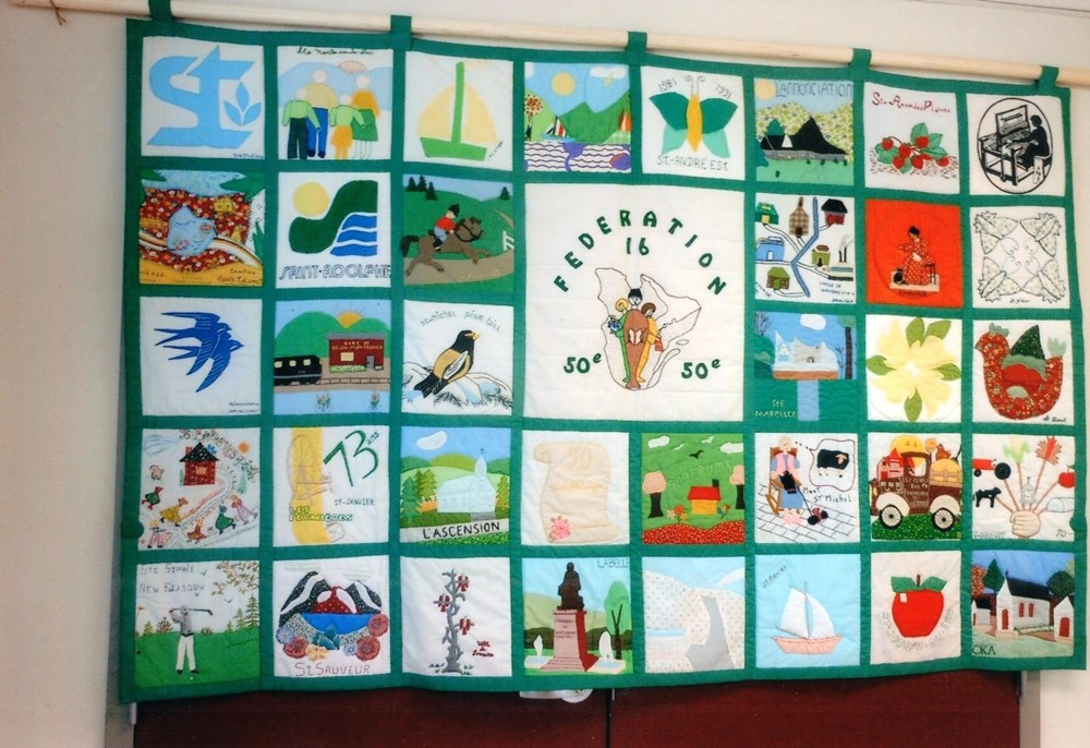 A quilt made up of nearly 40 squares. In the middle on a larger white square is the old logo of the Fermières showing three women in front of a map of Québec. It reads Fédération 16 (Federation 16) and 50e (50th anniversary). The surrounding squares are very colourful, and each one represents a different Cercle de Fermières.