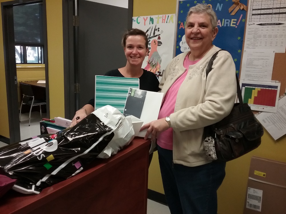 Two smiling women are standing side by side. One is holding a notebook and the other a sheaf of lined paper. On a desk in front of them is a full bag from the store Bureau Buro plus.