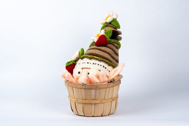 A crocheted gnome showing only his hat, head and fingers, with the rest hidden in a wooden basket, from which he seems to be emerging. He has big eyes and a prominent nose. On his brown striped hat there are flowers, strawberries, and a bug.