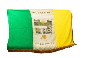 Drapeau vert, blanc et jaune à franges. Au centre, un champ, une femme ainsi qu’une ruche, un four à pain et une gerbe de blé sont peints à l’intérieur d’un blason. La devise des Fermières, « Pour la terre et le foyer », est inscrite au-dessus et en dessous du blason.