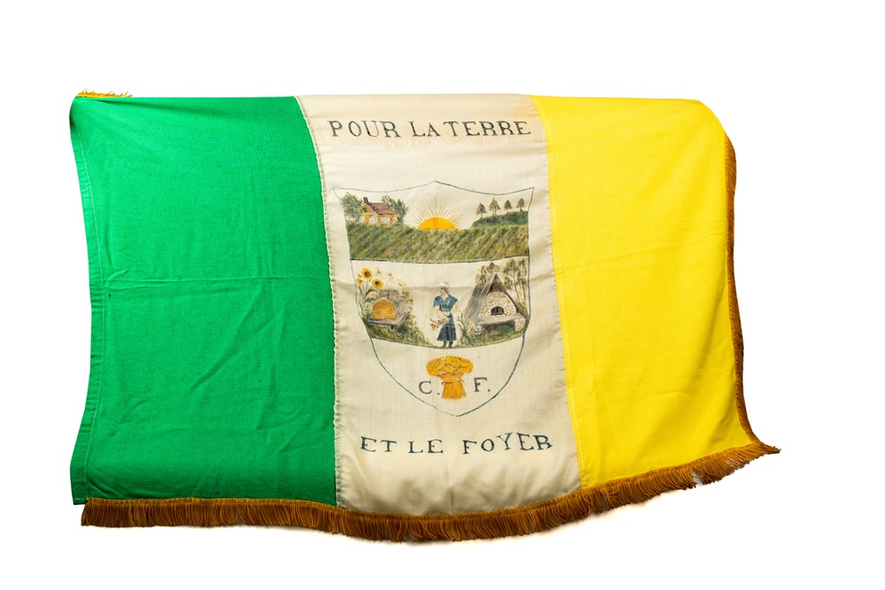 A green, white and yellow flag with a fringe along the bottom. In the middle, a field, a woman and a beehive, a bread oven and a sheaf of wheat are painted inside a shield. The Fermières’ motto, Pour la terre et le foyer (“For land and home”) is inscribed above and below the shield.