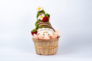 A crocheted gnome showing only his hat, head and fingers, with the rest hidden in a wooden basket, from which he seems to be emerging. He has big eyes and a prominent nose. On his brown striped hat there are flowers, strawberries, and a bug.