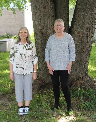 Two women are standing in front of a large tree in the summer. They are looking straight ahead and smiling.