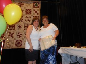 Two smiling women pose on a stage. In front of them a red balloon, a green balloon and a yellow balloon can be seen, and behind them hangs a red and burgundy quilt.