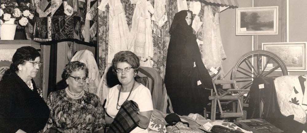 A black and white photo of three women inspecting a blanket. A quilt and some items of clothing are hanging behind them. On their right, blankets have been spread out on a table and a spinning wheel is displayed.