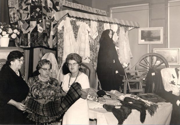 A black and white photo of three women inspecting a blanket. A quilt and some items of clothing are hanging behind them. On their right, blankets have been spread out on a table and a spinning wheel is displayed.