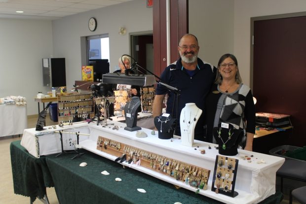 A smiling man and woman are standing behind a table on which earrings and necklaces are displayed.