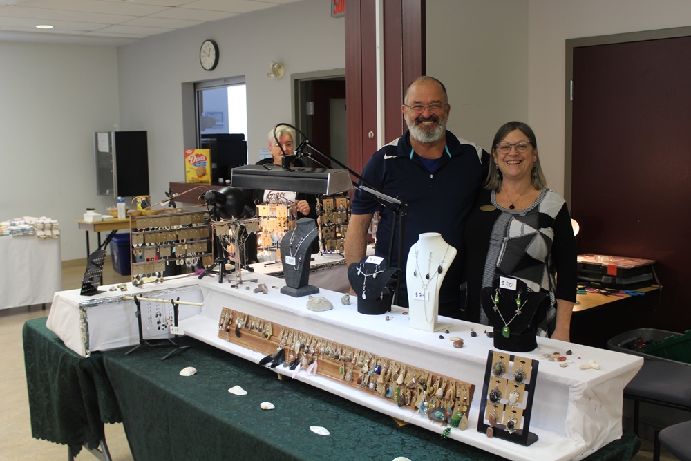 A smiling man and woman are standing behind a table on which earrings and necklaces are displayed.