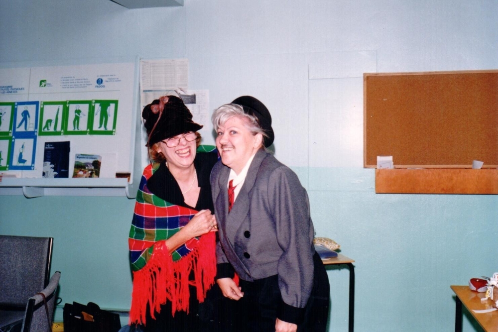 Two women in costumes are arm in arm, laughing. The one on the left is wearing a black hat and a red, blue and green shawl with a red fringe. The one on the right is wearing a hat, a grey jacket, a tie, and a false moustache.