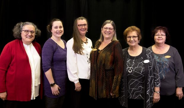 A photo of six smiling women posing next to one other.