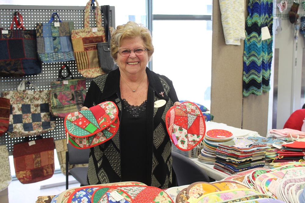 A smiling Fermière stands in her booth at the exhibition and sale, holding patchwork hot pads. Similar pieces are arranged on a table in front of her, and bags are hanging on a board behind her.