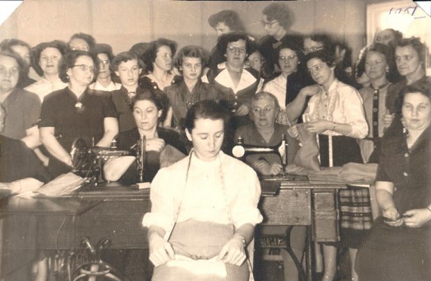 A black and white photo. Seated in the foreground is a woman wearing a white blouse with a measuring tape draped around her neck. Behind her are two womean with sewing machines in front of them. At the back, there are about twenty women standing.