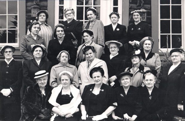 A black and white photograph taken in front of a building, showing twenty-one women in four rows, each wearing a jacket or coat and a hat. The women in the first row are seated while the others are standing.