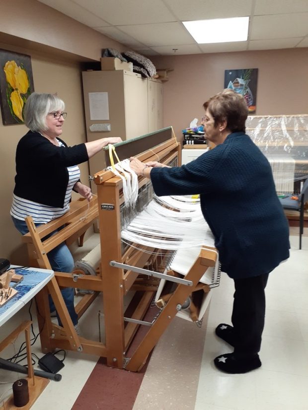 Two women are standing on either side of a wooden loom. They are in the process of setting up the loom with white threads. Another loom can be seen in the background.