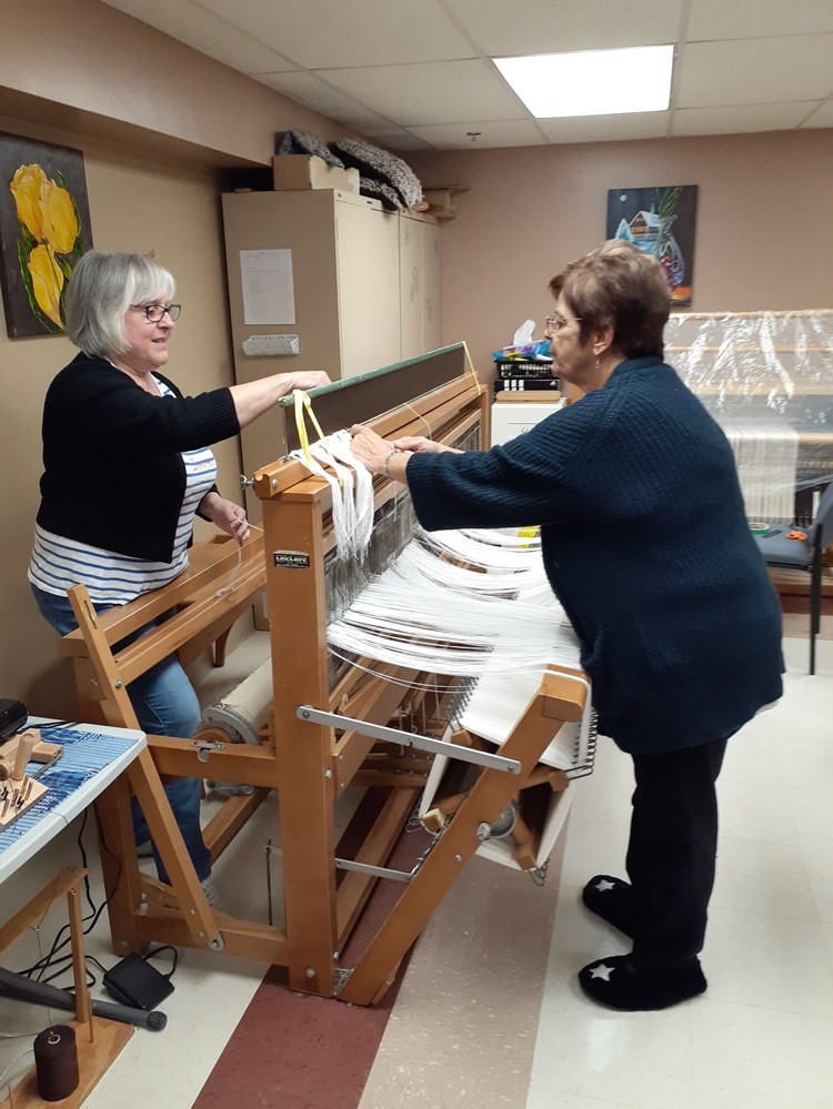 Deux femmes sont debout chacune d’un côté d’un métier à tisser de bois. Elles procèdent au montage du métier avec des fils blancs. À l’arrière-plan, on aperçoit un autre métier à tisser. 