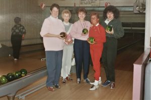 Five women wearing bowling shoes are standing in a bowling alley. They hold a ball in their hands and are smiling.