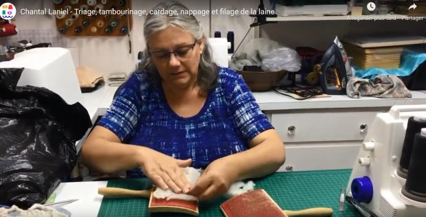 A woman with grey hair wearing a blue shirt sits behind a table. A cutting mat is on the table. On the cutting mat, there are two hand carders. With her hands, the woman places white wool on one of the carders.