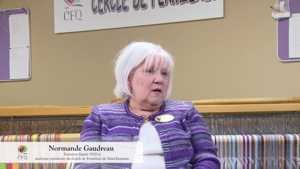 A white hair woman wearing a striped violet cardingan is sitting in front of a loom.