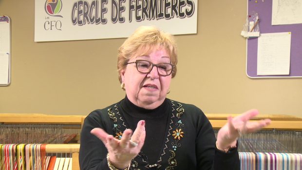 Close-up of a woman with short blond hair wearing a black top. She moves her hands while she speaks. The woman is in front of a loom.