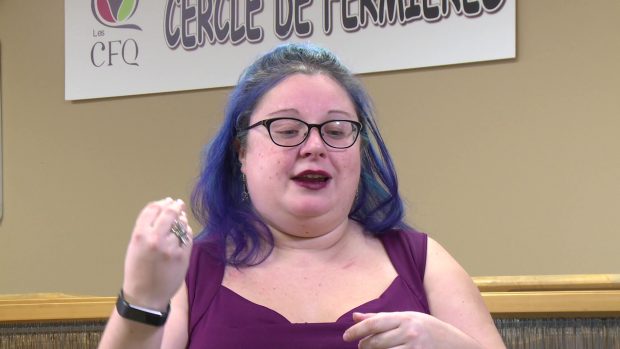 Close-up of a woman with blue hair wearing a purple top. She is explaining something and she is moving her hands. Behind her, we can see the upper part of a loom.