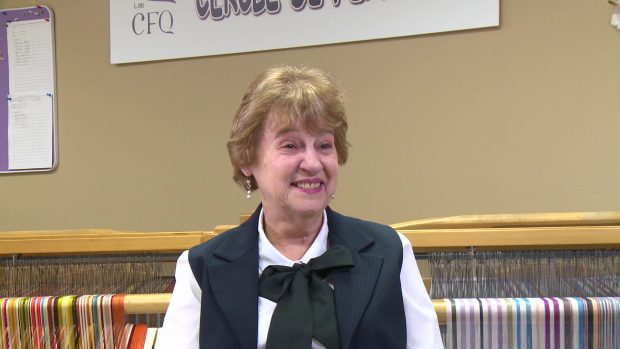 Close-up of a smiling woman with short blond hair. She wears a white blouse, a blue sleeveless jacket and a black bow. She is in front of a loom.