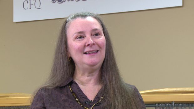 Close-up of a woman with long grey hair. She wears a brown blouse and a gold necklace. Behind her, we can see the upper part of a loom.