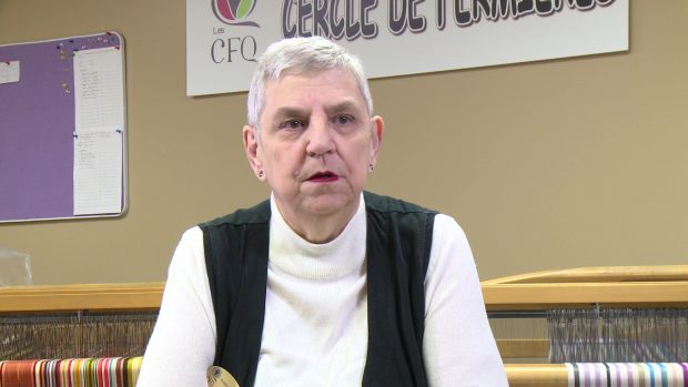 Close-up of a women with very short white hair. She wears a white turtleneck and a black sleeveless jacket. Behind her, there is a loom.