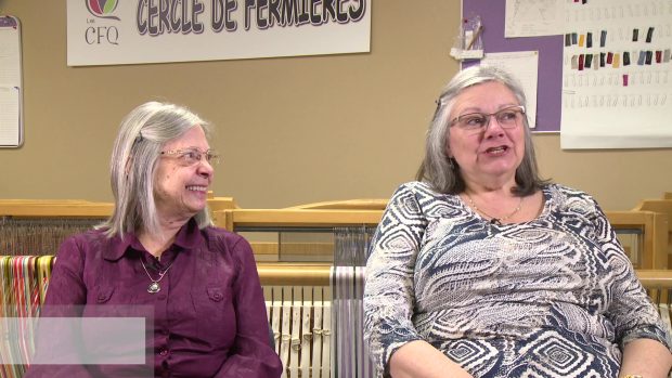 Two women with grey hair are sitting, side by side, in front of a loom. The one on the left watches the one on the right, smiling. The one on the right speaks.