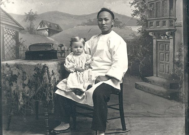 Chinese domestic worker sitting in a chair posing for a photograph with a young child on his right knee.