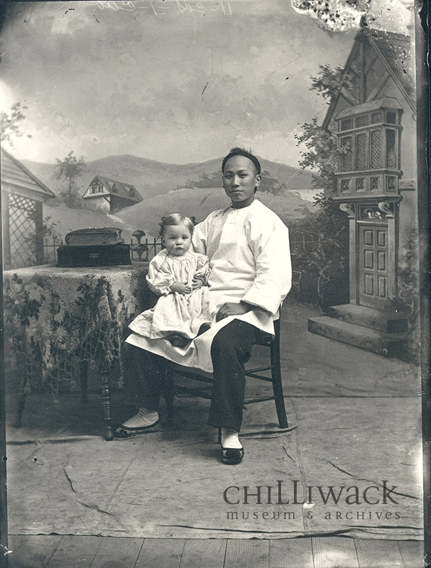 Chinese domestic worker sitting in a chair posing for a photograph with a young child on his right knee.