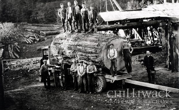 Group portrait of thirteen men standing with fir log loaded on truck at the Orion Bowman mill