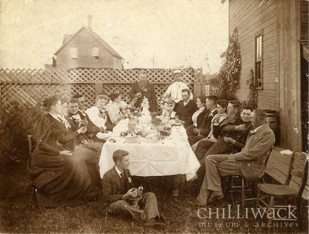 Group portrait of eighteen individuals around a table with one individual sitting on the ground. House and fence visible in background.