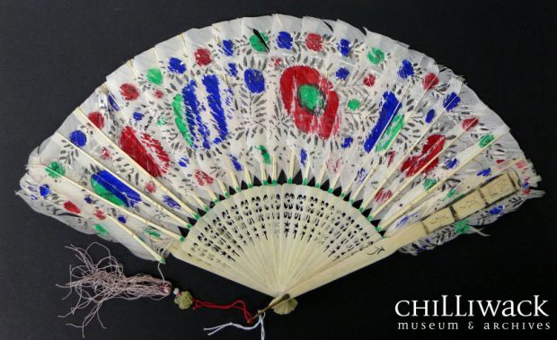 Painted white fan with red, green and blue floral design containing parrots. Light purple tassel hanging from brass loop