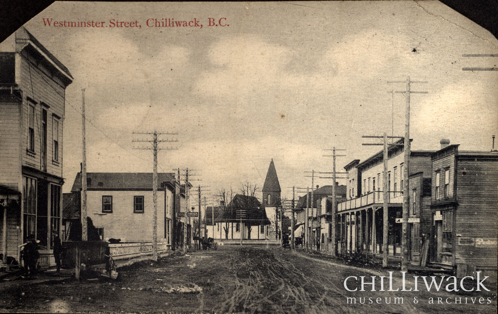 Black and white postcard of Westminster Street in Chilliwack, British Columbia. Street lined with two story buildings with church in background.