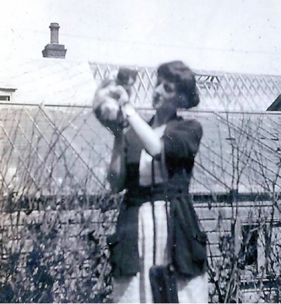 Black and white picture. Of women holding a cat she is standing in front of two glass roofed buildings.