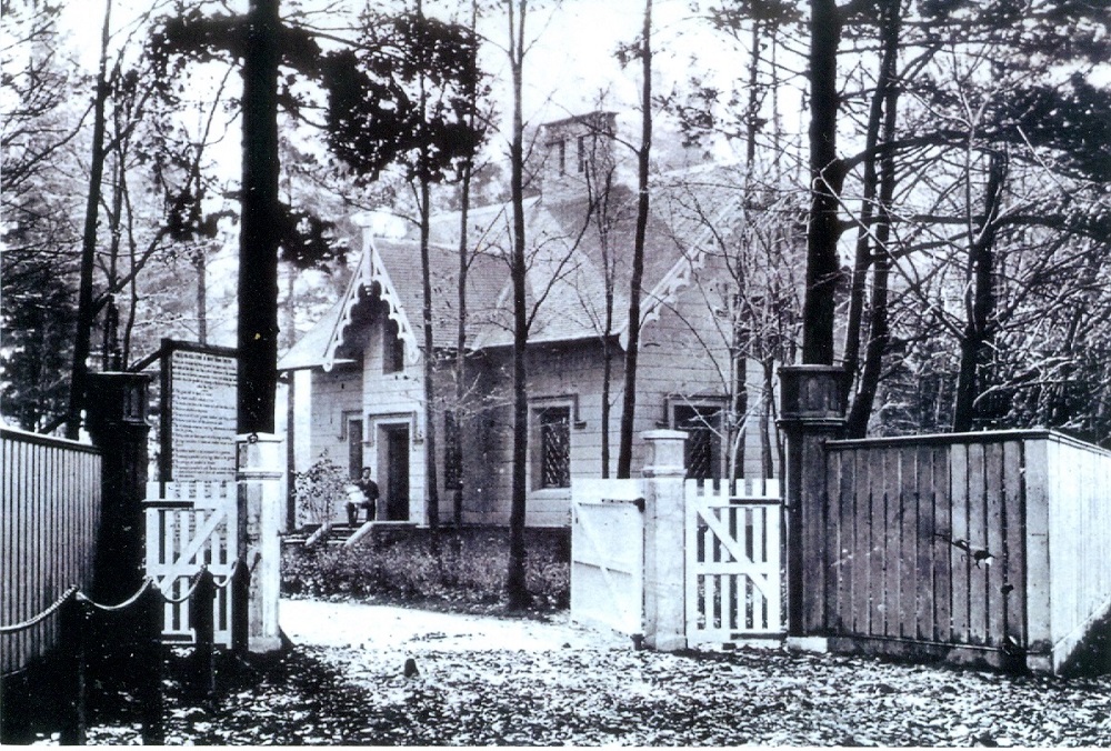 Black and white picture of a house in the woods. A man is sitting on the front step holding a white-clothed baby. There is also a large wood fence in front of the house.