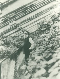 Black and white picture of a man standing in a room with lots of plant. The room has a slanted ceilling. The man is dressed in dark pant, a dark vest and a white shirt. He has a long white beard and is wearing a circular hat.