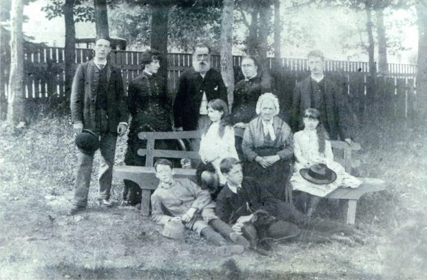 Black and white photo of a family in a courtyard surrounded by trees and a wooden fence. In this picture, there are ten people. Five people are standing, three are sitting on a bench, and two are lying on the ground in the grass. They dress all their best Sunday.