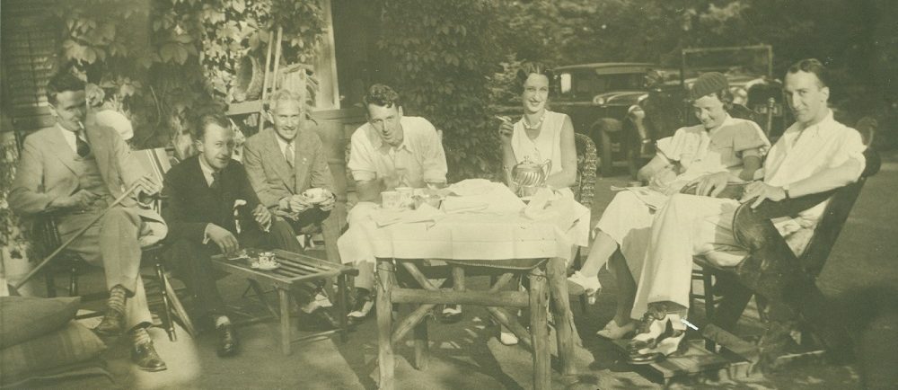 Sepia picture of people sitting in chairs around a tea set on a table covered in white linen