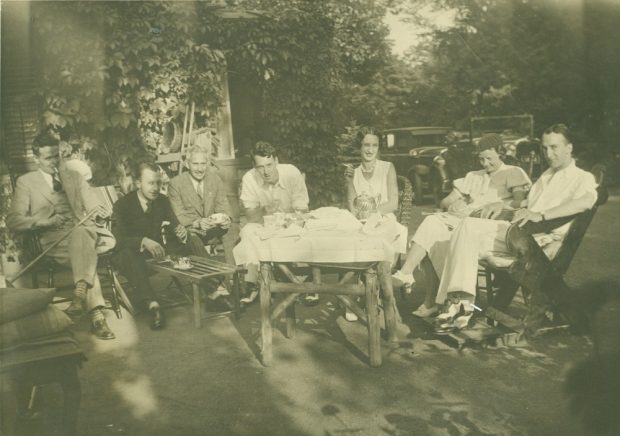 Sepia picture of people sitting in chairs around a tea set on a table covered in white linen